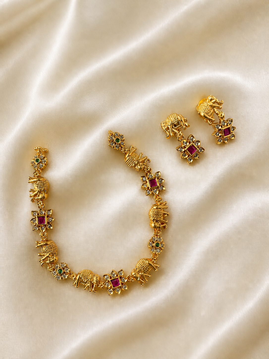 A traditional gold-plated link necklace and matching drop earrings laid out on a cream satin fabric, featuring alternating motifs of detailed carved elephants and square-cut ruby red stones enclosed in a floral star border.