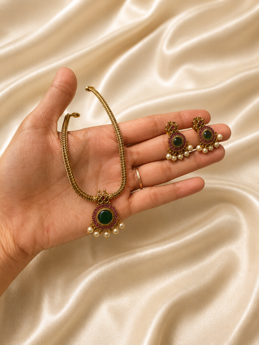 A delicate traditional jewelry set displayed on white satin fabric, featuring a gold-plated short necklace with linked leaf and flower motifs embedded with small pinkish-red kemp stones, paired with matching dangling earrings.