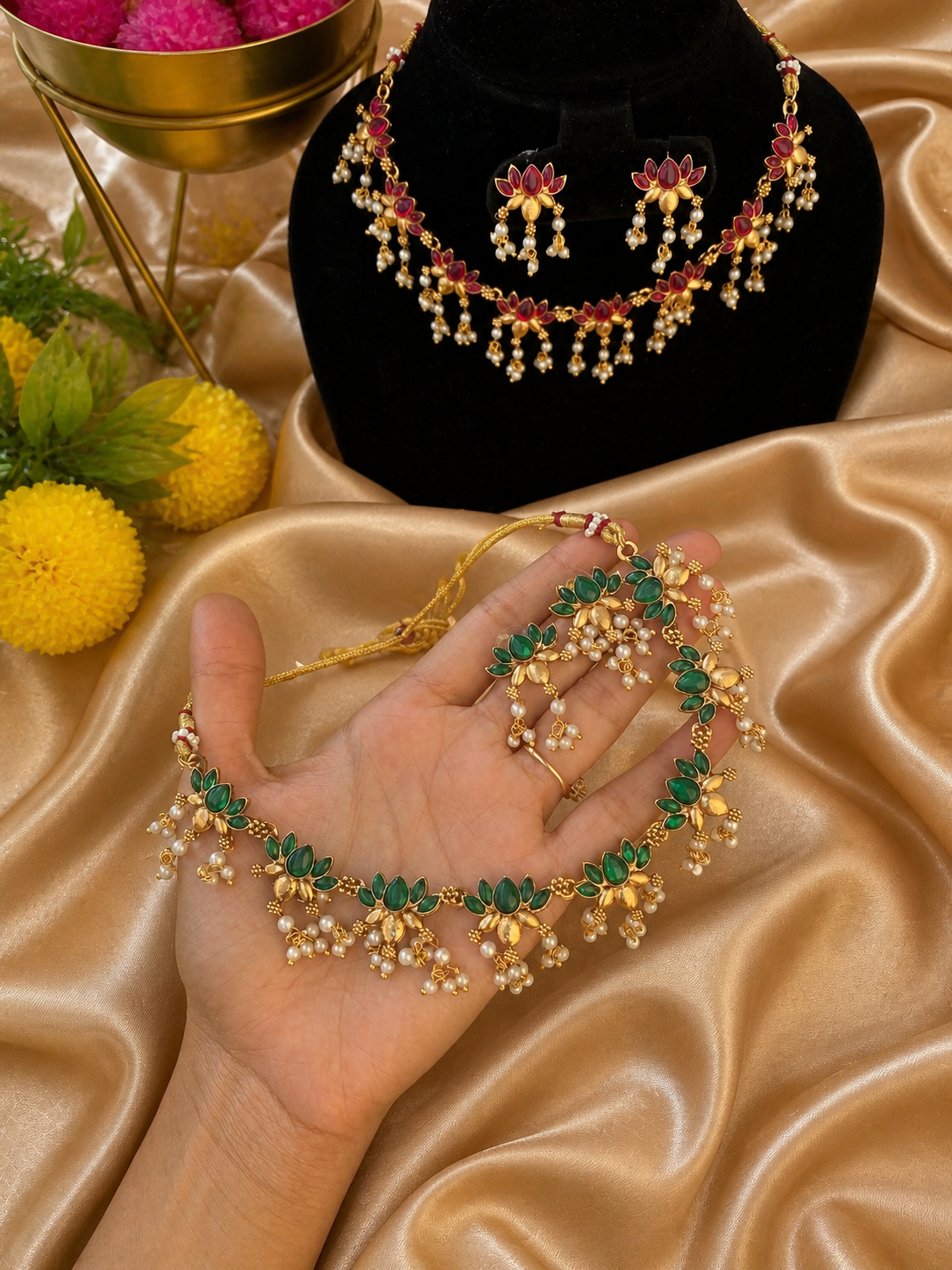 A traditional South Indian jewelry set showcasing a red kemp stone necklace on a black display bust and a matching green kemp stone necklace held in a hand. Both gold-plated necklaces feature pointed leafy gem motifs and clustered pearl drops over a golden fabric background.