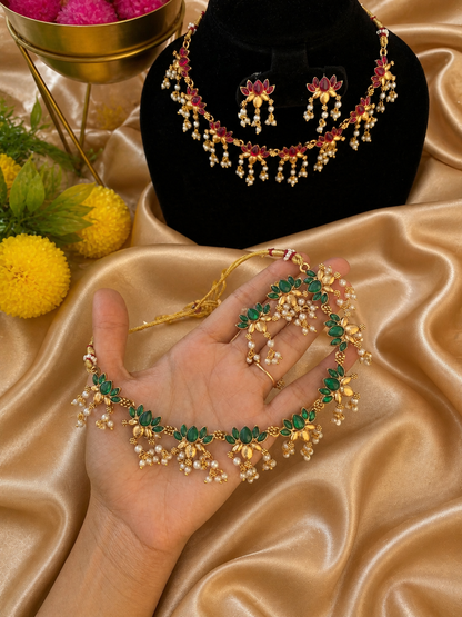 A traditional South Indian jewelry set showcasing a red kemp stone necklace on a black display bust and a matching green kemp stone necklace held in a hand. Both gold-plated necklaces feature pointed leafy gem motifs and clustered pearl drops over a golden fabric background.