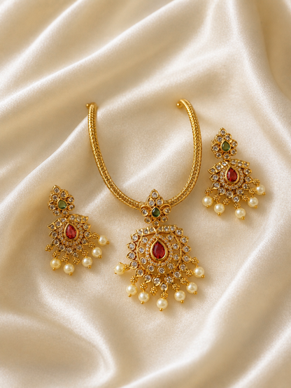 An elegant traditional jewelry set laid out on ivory satin fabric, featuring a thick woven gold-plated chain with a large, ornate teardrop-shaped pendant embedded with a central red ruby stone, surrounded by clear crystals and a fringe of white pearls, complete with matching dangling statement earrings.