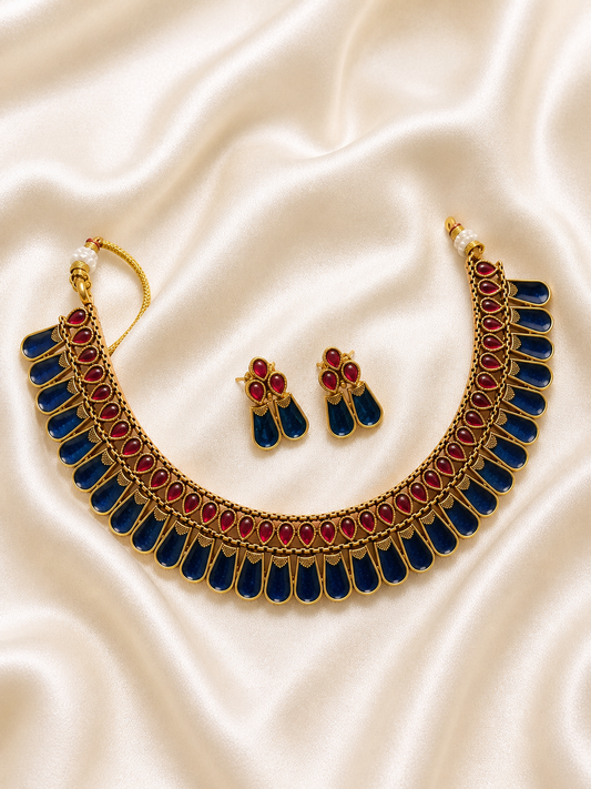 A striking antique gold-plated collar necklace and matching drop earrings laid out on a cream satin fabric, featuring a structured crescent design adorned with a row of teardrop ruby red kemp stones fringed by elongated panels of royal blue enamel work.