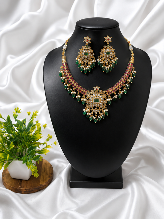 A stunning antique gold-plated festive necklace held in a hand over cream satin fabric, featuring a curved collar structure lined with small pink kemp stones, a large central statement pendant featuring a square emerald surrounded by white CZ petals, and a colorful fringe of hanging emerald green beads and tiny pearls.
