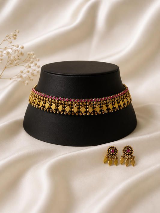 traditional antique gold-plated choker sets on ivory satin. The set features a band of small pink stones, intricate gold beadwork, and hanging leaf-shaped charms, paired with matching pink-stone stud earrings with leaf drops.