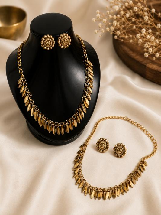 Antique gold-plated jewelry set featuring a link chain necklace adorned with a row of dangling, textured leaf-shaped charms. One set is displayed on a black glossy bust stand with matching circular floral-patterned stud earrings, while an identical set lies flat on a cream satin cloth