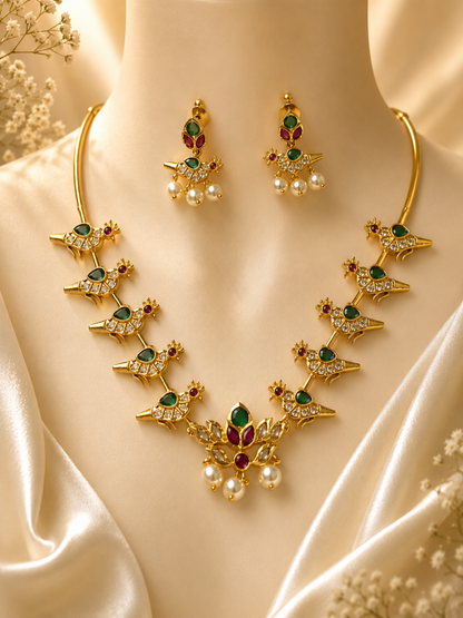A traditional gold-plated collar necklace featuring a series of stylized parrot motifs embedded with green, red, and white crystals, leading to a central floral pendant with hanging pearls. The set includes matching parrot-shaped drop earrings and is displayed on a cream-colored bust.