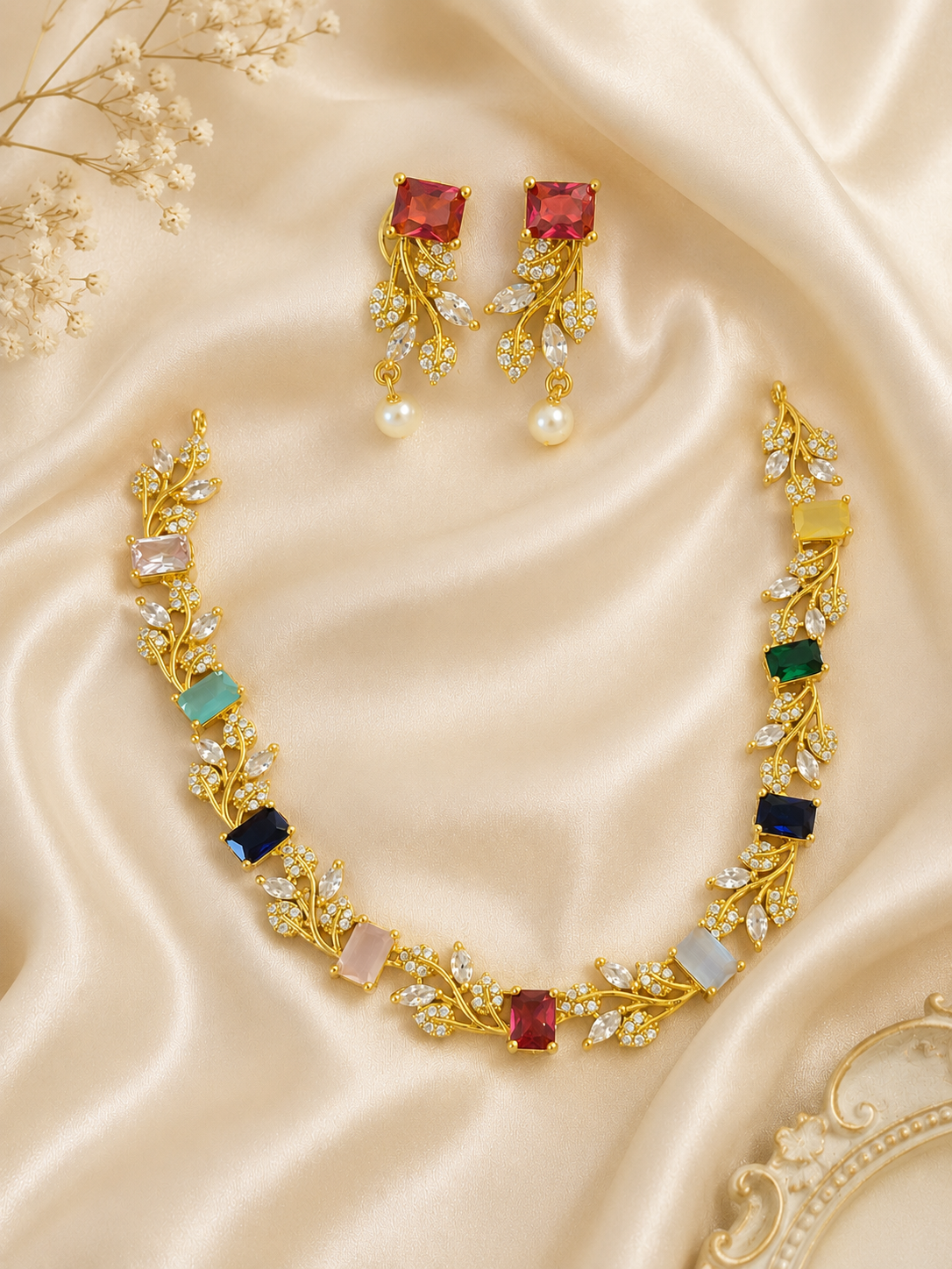 Vibrant gold-plated jewelry set featuring a flexible vine-style necklace intertwined with small clear crystals and larger, multi-colored rectangular gemstones in shades of red, blue, green, and pink. The set includes matching long drop earrings with a large red top stud and a hanging pearl, all arranged flat on champagne satin.