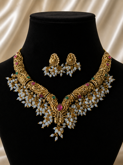 Traditional South Indian antique gold guttapusalu choker necklace featuring an intricate curved gold band studded with pink ruby and green emerald stones, densely fringed with clusters of small white seed pearls. The set is accompanied by matching pearl-fringed stud earrings, presented flat on smooth champagne satin.