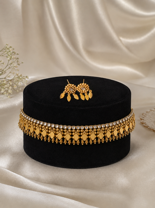 Traditional antique gold-plated elakkathali choker necklaces arranged horizontally on ivory satin. Choker features a row of small clear white stones, intricate gold beadwork, and hanging leaf-shaped charms, paired with matching cluster stud earrings with leaf drops.