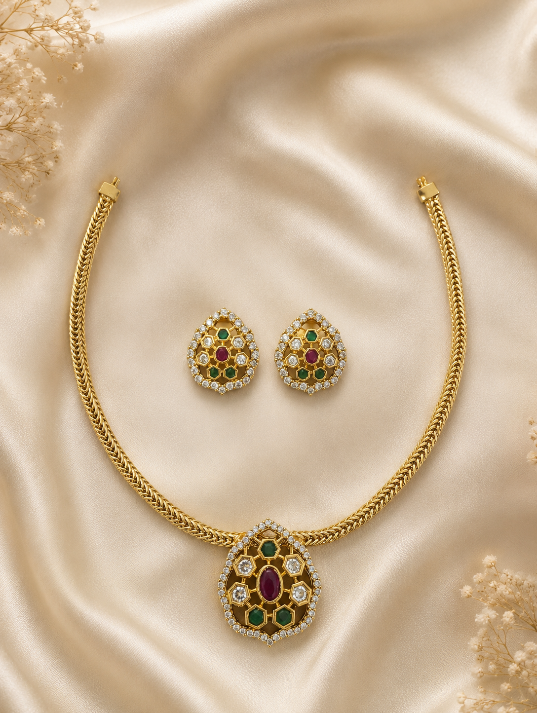 Latha heritage jewelry set featuring a gold-toned braided chain necklace with a large teardrop-shaped kemp pendant containing a center ruby stone, emerald accents, and clear crystal framing, laid flat on champagne satin silk alongside matching teardrop stud earrings.