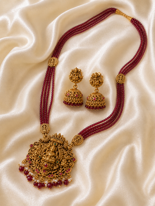 Antique gold temple jewelry set featuring a multi-strand maroon beaded long necklace with a large, intricate Goddess Lakshmi pendant and matching golden jhumka earrings, laid flat on a shimmering champagne satin fabric.