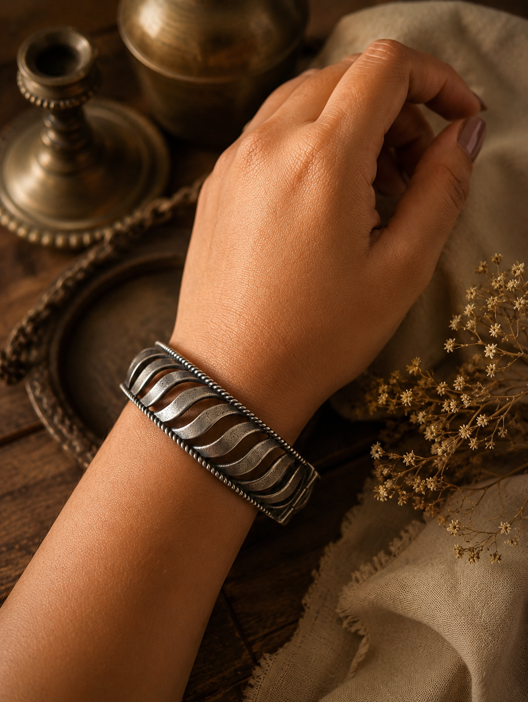A close-up shot of a person's wrist wearing an oxidized silver cuff bracelet with cut-out wave patterns and twisted border detailing, set against a warm, rustic background with vintage brass items =