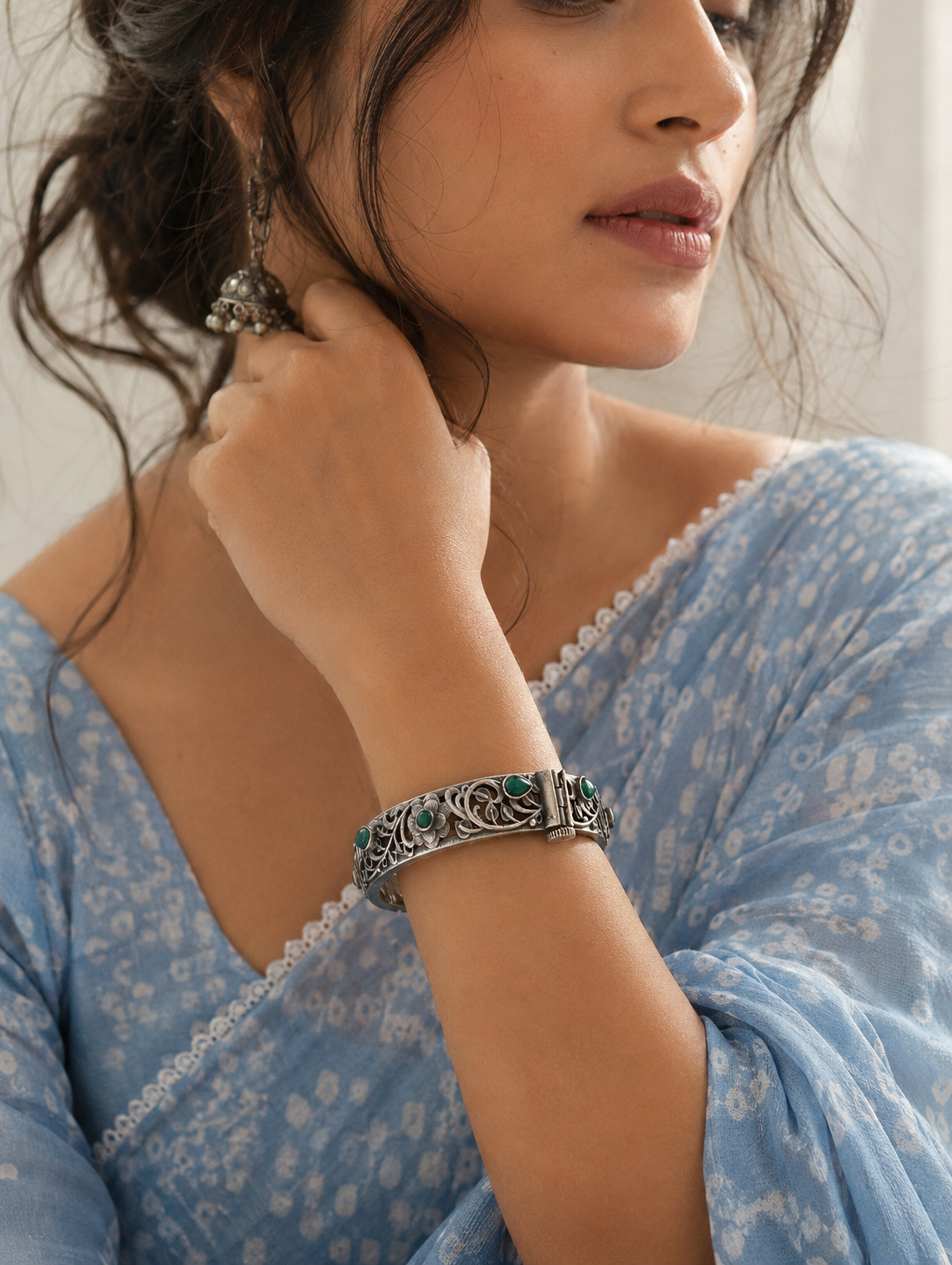 Woman wearing a blue floral dress and silver bracelet with green stones, adjusting her hair.