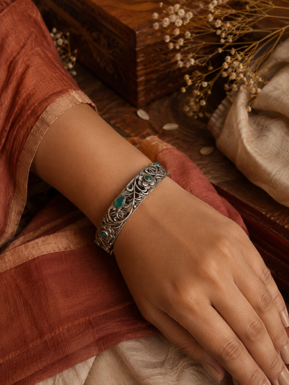 A high-angle shot of a person’s wrist wearing an intricate oxidized silver filigree cuff bracelet adorned with green stones and carved flower motifs, set against a warm aesthetic background with a wooden box and a terracotta saree