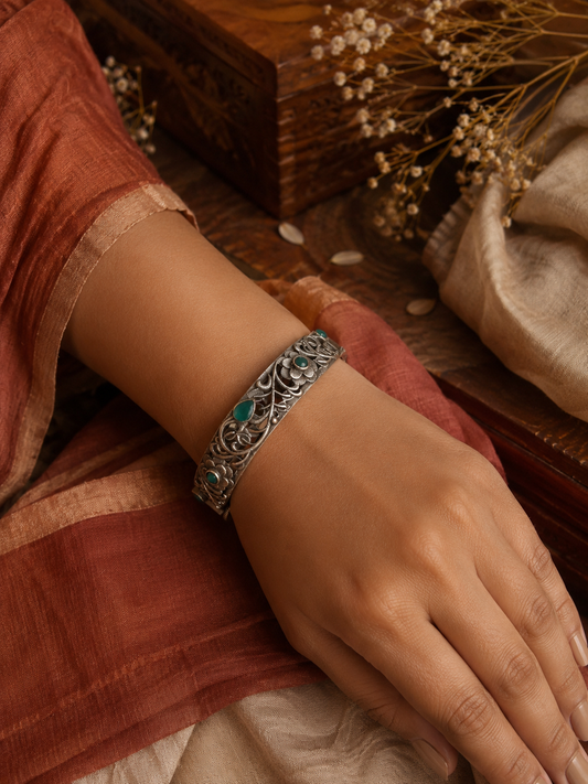 A high-angle shot of a person’s wrist wearing an intricate oxidized silver filigree cuff bracelet adorned with green stones and carved flower motifs, set against a warm aesthetic background with a wooden box and a terracotta saree