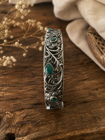 Silver bracelet with green stones on a wooden surface with dried flowers and fabric.