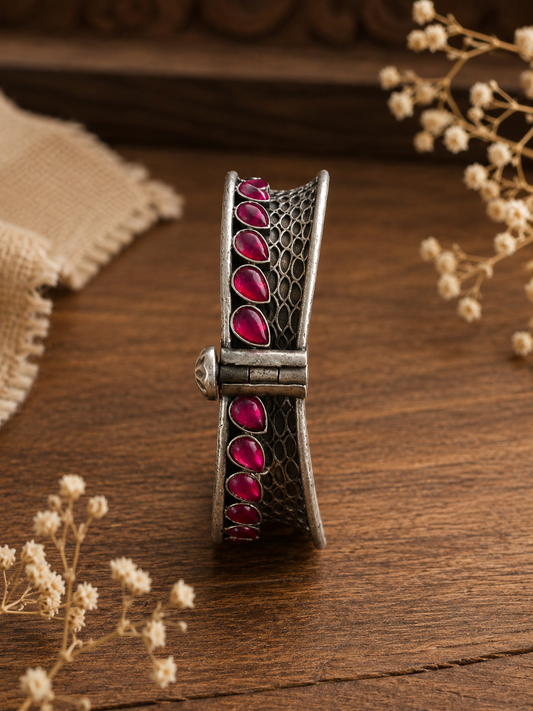A side-profile shot of a thick oxidized silver kada bangle standing vertically on a wooden table, showcasing a row of teardrop-shaped ruby pink gemstones, intricate dark mesh texturing, and a vintage screw-lock mechanism 