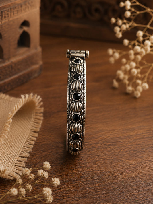 A side-profile shot of a slim oxidized silver kada standing upright on a dark wooden table, showcasing a continuous pattern of round faceted black stones alternating with ribbed silver beads