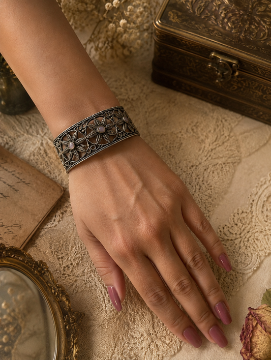 A detailed overhead shot of a person's wrist wearing a wide oxidized silver cuff bracelet with geometric floral jali cutouts and small light pink stone centers, set against an antique layout with lace, a brass box, and a handheld mirror 