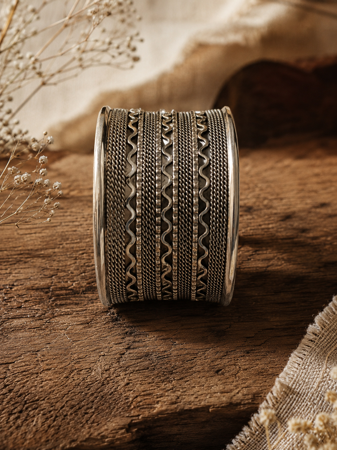 A front-facing shot of a wide, multi-layered oxidized silver cuff bangle standing vertically on a rustic dark wooden surface, showcasing detailed horizontal patterns of twisted rope borders, dark mesh lines, and wavy metallic accents 