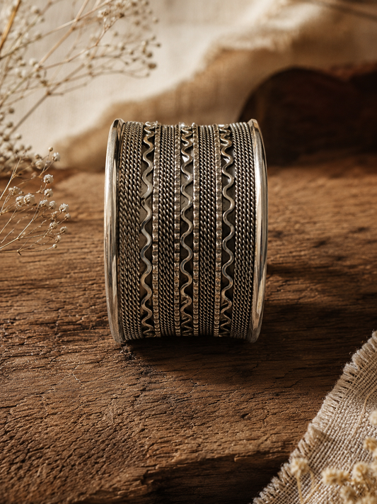 A front-facing shot of a wide, multi-layered oxidized silver cuff bangle standing vertically on a rustic dark wooden surface, showcasing detailed horizontal patterns of twisted rope borders, dark mesh lines, and wavy metallic accents 