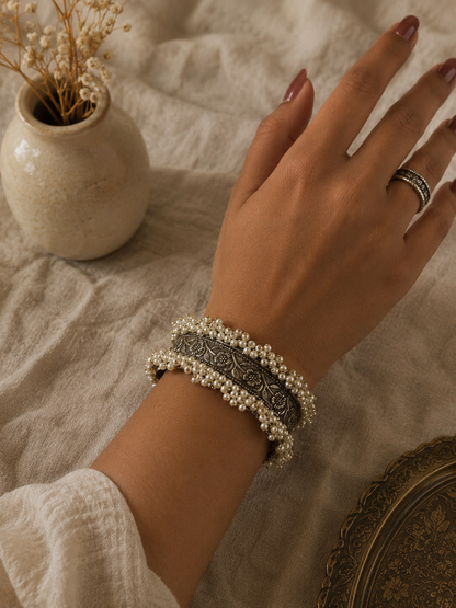 A close-up of a model's wrist wearing the Kalpana oxidized silver kada bangle, detailed with fine floral engraving and small white pearls along the borders, set against a neutral fabric background.