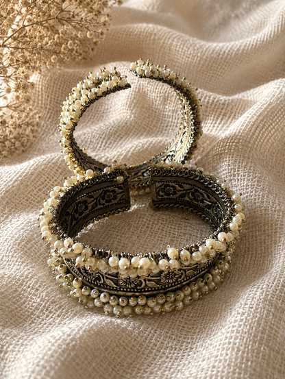 A pair of Kalpana oxidized silver open-style kada bangles resting on a textured cream fabric, showcasing detailed floral metalwork and dense borders of small white pearl beads.