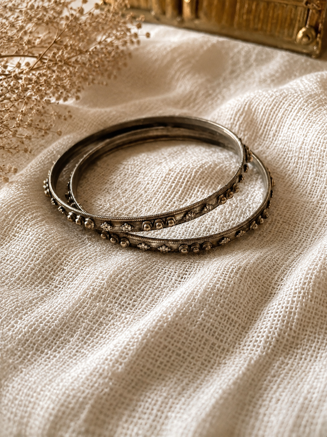 A pair of Rama slim oxidized silver bangles with raised metallic bead and flower motifs, displayed on a textured white linen fabric under warm lighting.