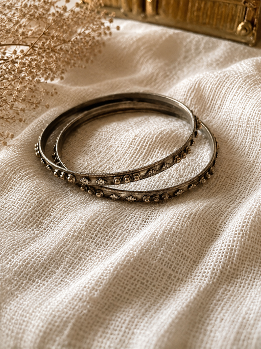 A pair of Rama slim oxidized silver bangles with raised metallic bead and flower motifs, displayed on a textured white linen fabric under warm lighting.