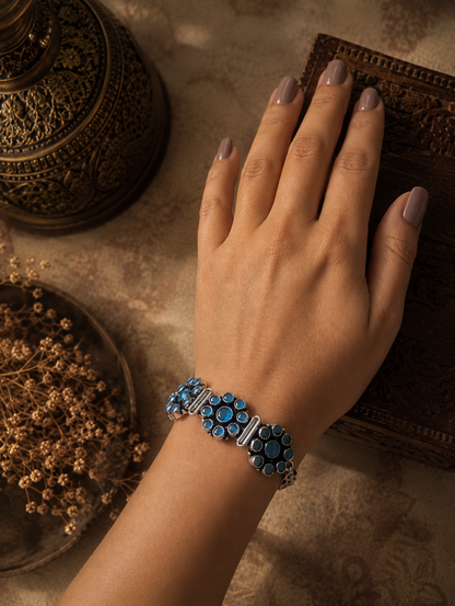  Kalavathi oxidized silver bracelet featuring floral stone-studded patterns in blue, displayed on a textured fabric background.