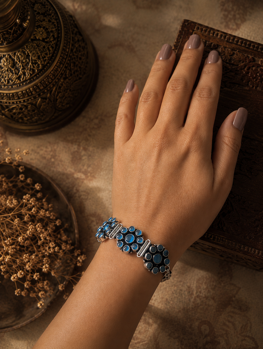  Kalavathi oxidized silver bracelet featuring floral stone-studded patterns in blue, displayed on a textured fabric background.