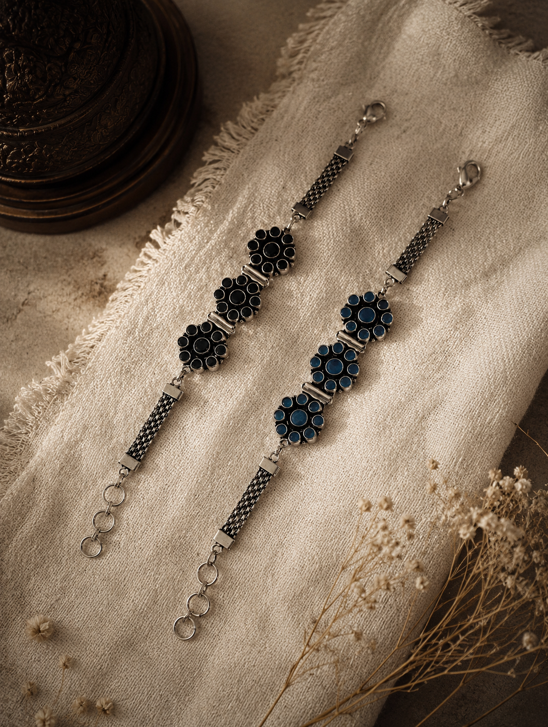Two Kalavathi oxidized silver bracelets featuring floral stone-studded patterns in black and deep blue, displayed on a textured fabric background.