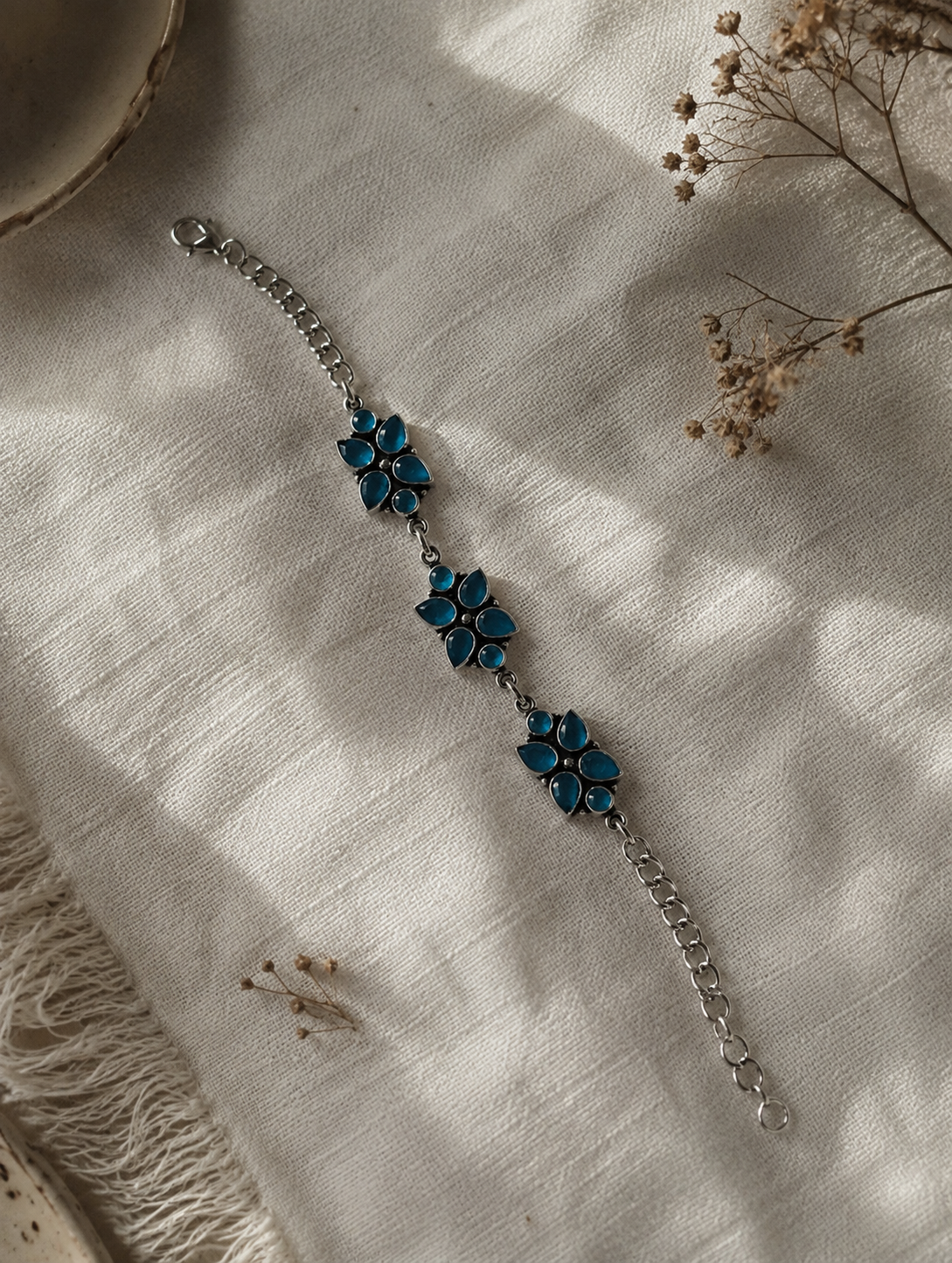 An oxidized silver link bracelet featuring three floral clusters made of teardrop-shaped blue stones, displayed on a textured white linen fabric under soft sunlight.