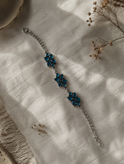 An oxidized silver link bracelet featuring three floral clusters made of teardrop-shaped blue stones, displayed on a textured white linen fabric under soft sunlight.
