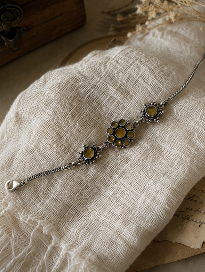 The Sharmila oxidized silver bracelet featuring a center floral cluster and flanking circular charms set with honey-yellow stones, arranged neatly on a crumpled off-white linen cloth.