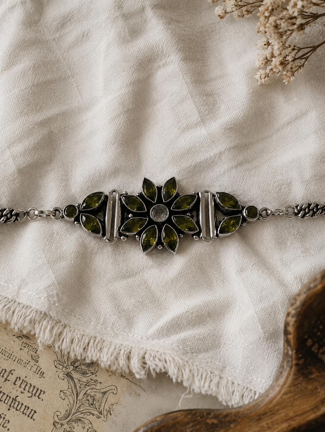 An oxidized silver statement bracelet showcasing a large central flower motif and side accents crafted from olive green marquise-cut crystals, resting on a textured off-white linen cloth.