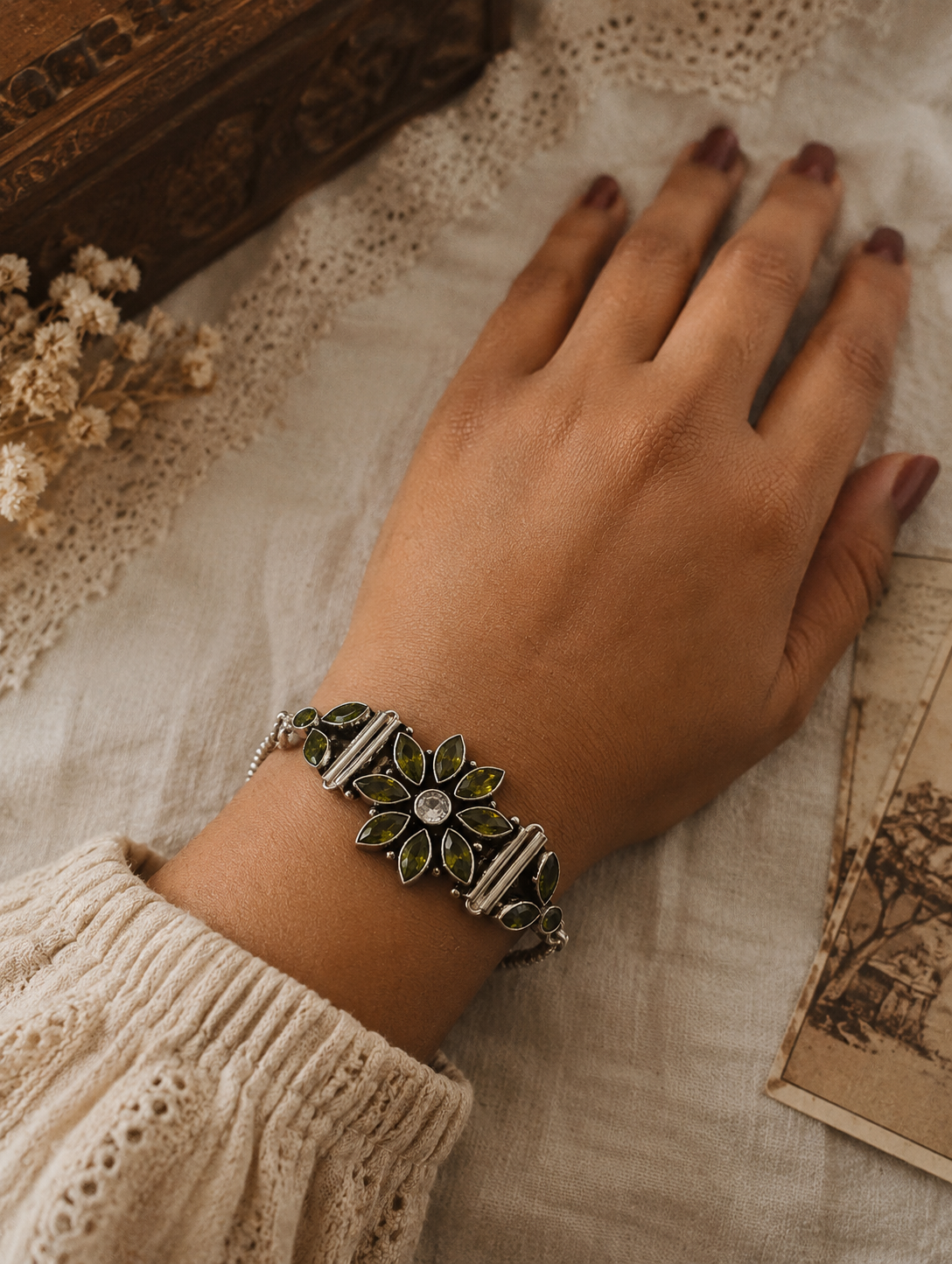 Hand wearing a decorative bracelet on a textured surface with a wooden box and dried flowers in the background.