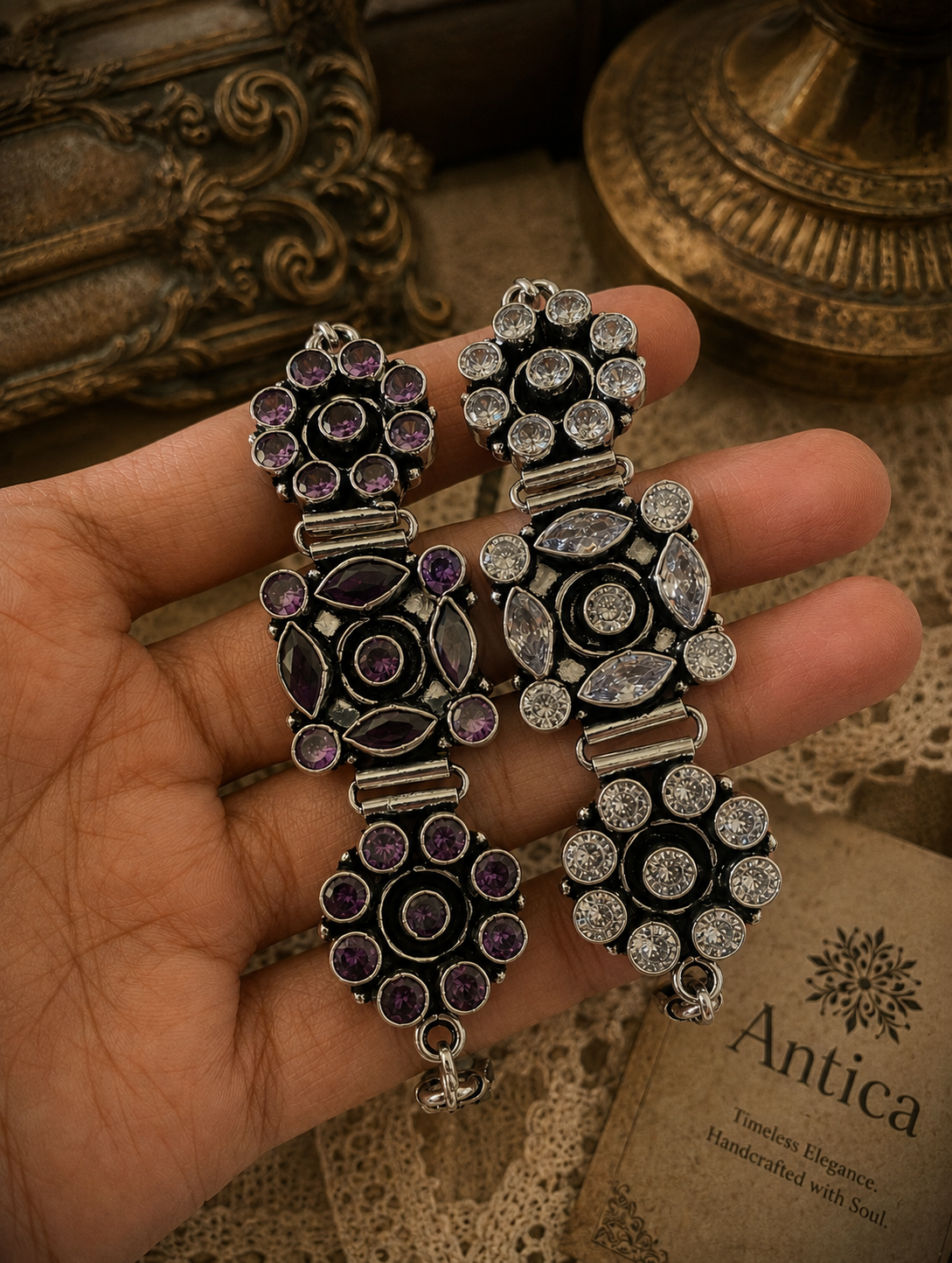 A person holding two Nazaakat statement bracelets crafted from oxidized silver; one is set with deep purple crystals and the other with clear crystals, featuring intricate floral and marquise-cut patterns.
