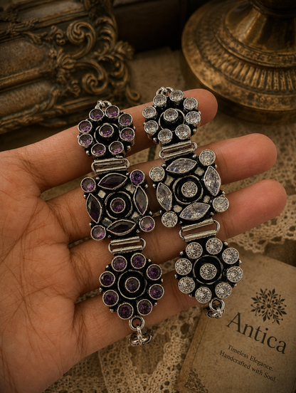 A person holding two Nazaakat statement bracelets crafted from oxidized silver; one is set with deep purple crystals and the other with clear crystals, featuring intricate floral and marquise-cut patterns.