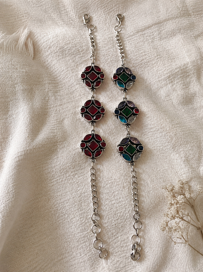 Two Ashwathi oxidized silver link bracelets laid side-by-side on a white fabric background; one features monochromatic ruby-red geometric stones, and the other showcases a multi-colored mosaic of green, blue, purple, and red stones.