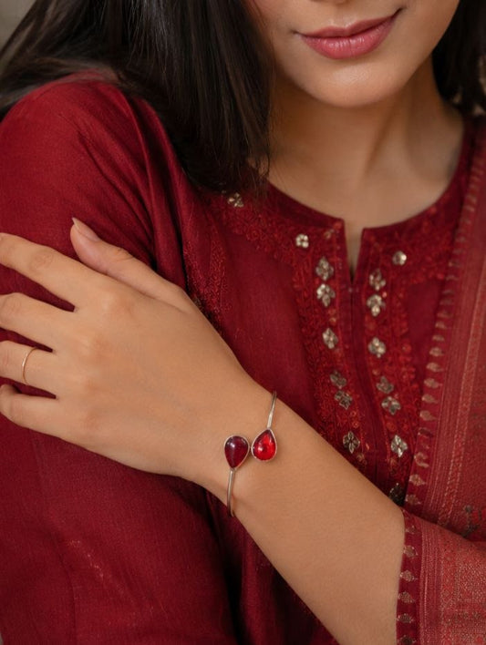 A model wearing a minimalist silver open-cuff bracelet featuring two vibrant teardrop-shaped ruby red stones on the wrist, paired with a matching deep red embroidered outfit.