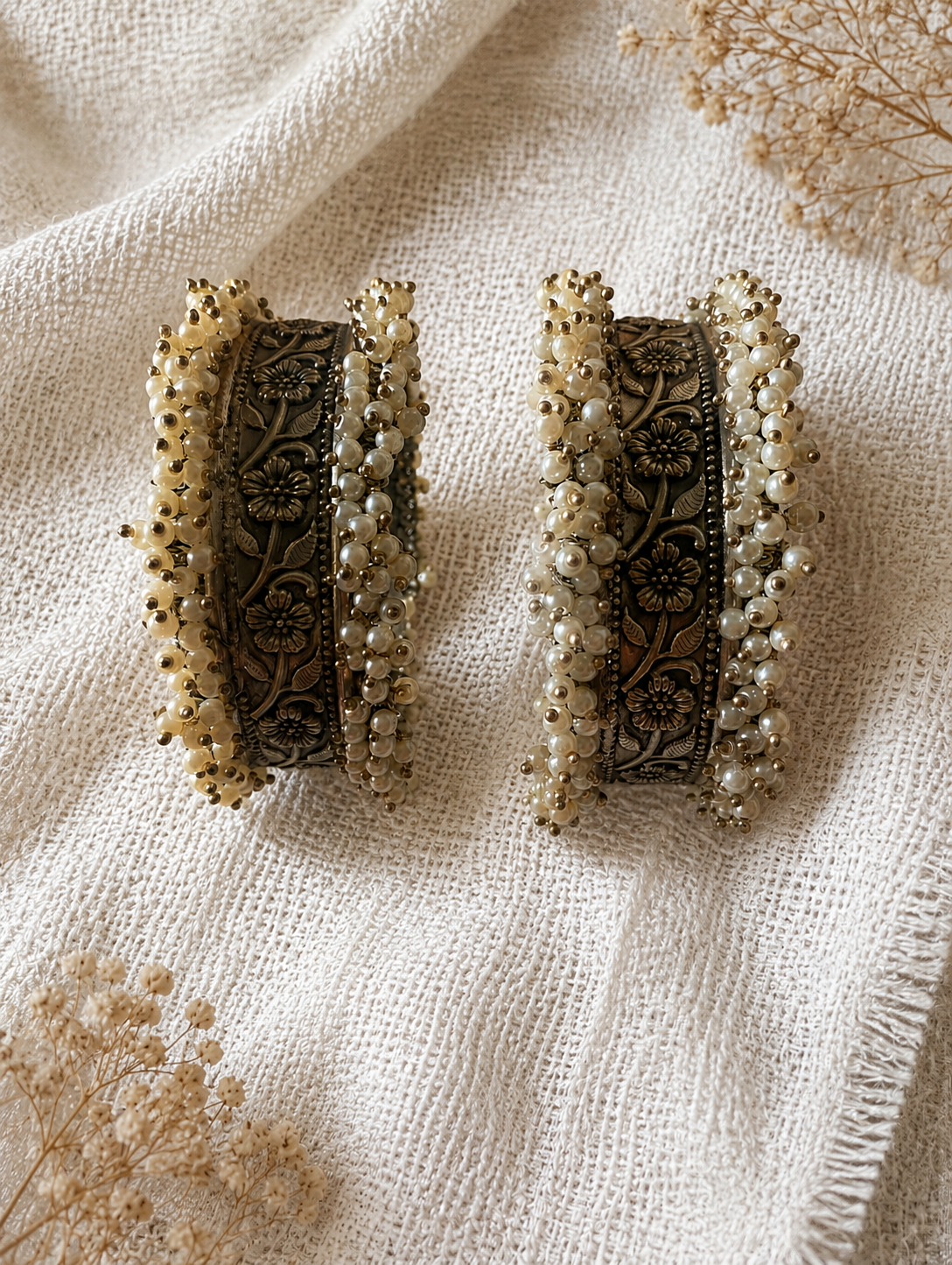 A pair of heavy Kalpana oxidized silver open-style kada bangles embellished with rows of small white pearls along the outer borders and detailed floral metal engravings inside, displayed on textured cream fabric.