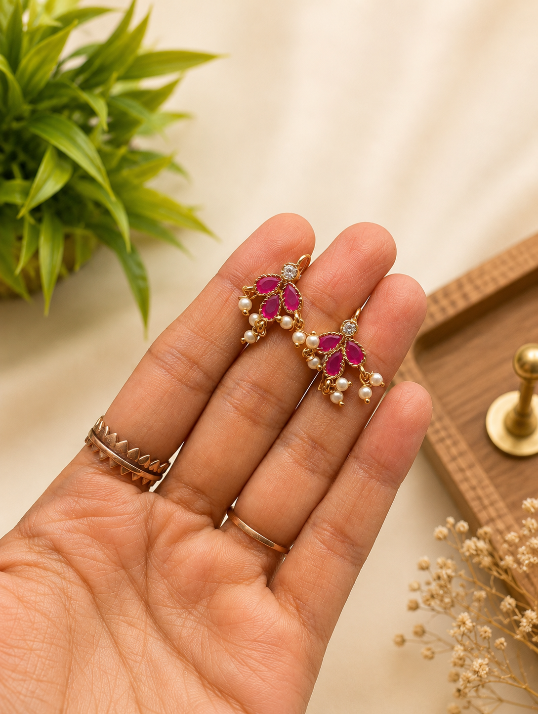 Pair of traditional gold-plated Maharashtrian Bugudi helix earrings adorned with leaf-shaped pink ruby stones and tiny dangling pearls, held in a hand.