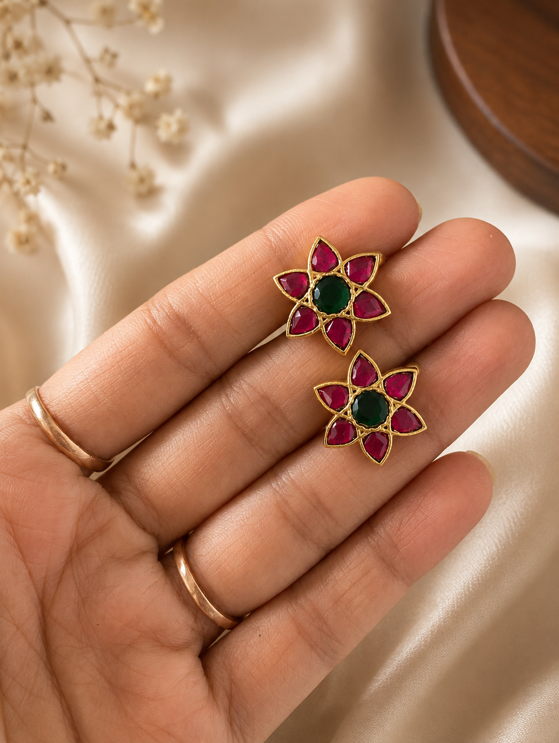 Pair of traditional gold-plated floral-shaped Bugudi helix ear cuffs featuring a round green center stone and pink-ruby petals, held in a hand.