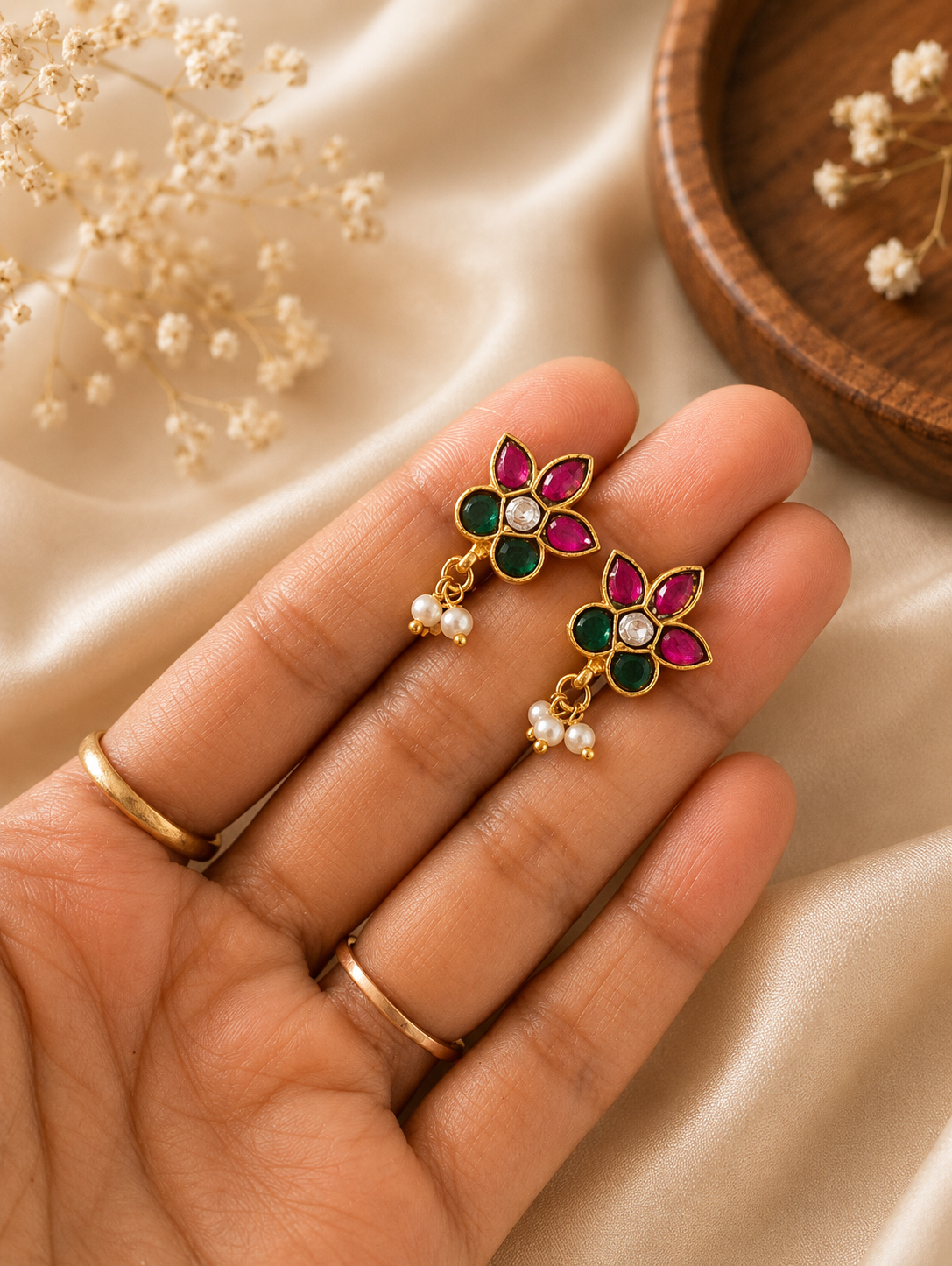 Close-up shot of a pair of gold floral ear cuffs featuring pink and green kemp stones with tiny hanging white pearls, held in a person's hand against a soft silk background.