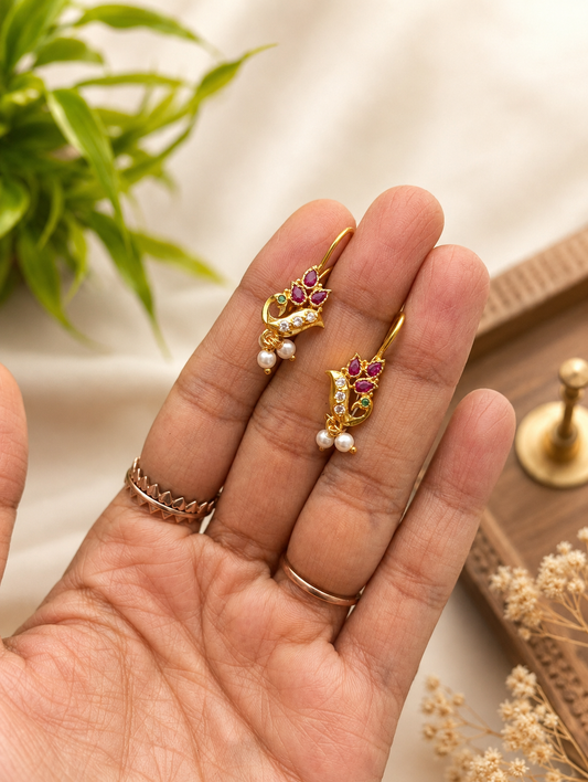 A pair of traditional gold Peacock style Bugudi earrings held in a hand, featuring intricate peacock designs adorned with pink ruby stones, white stones, small green accents, and dangling white pearls.