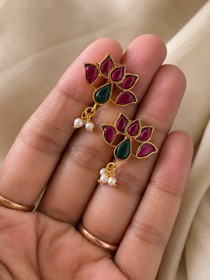 Close-up of a pair of gold-plated Lotus Jadau Kundan Bugudi earrings held in a hand, featuring a lotus-shaped design set with pink stones, a teardrop green stone center, and tiny dangling white pearls.