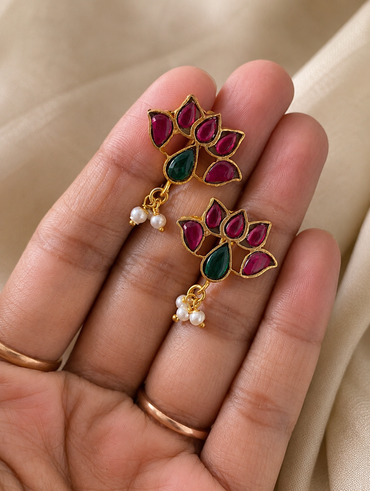 Close-up of a pair of gold-plated Lotus Jadau Kundan Bugudi earrings held in a hand, featuring a lotus-shaped design set with pink stones, a teardrop green stone center, and tiny dangling white pearls.