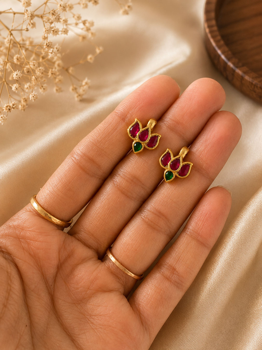 Close-up view of a pair of small, gold-plated mini lotus bugudi earrings resting on a hand, featuring three pink stones forming the petals and a small teardrop green stone at the base.