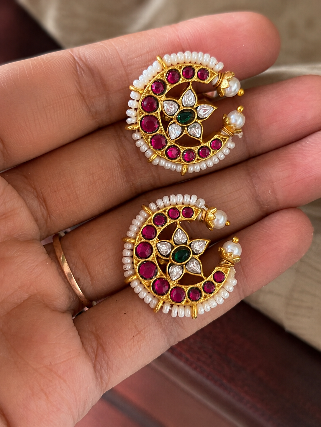 Close-up of a pair of traditional South Indian Chandra Bugudi gold ear cuffs held in a hand, featuring a crescent moon shape lined with tiny white pearls, embedded rubies, and a central star-shaped stone flower motif.